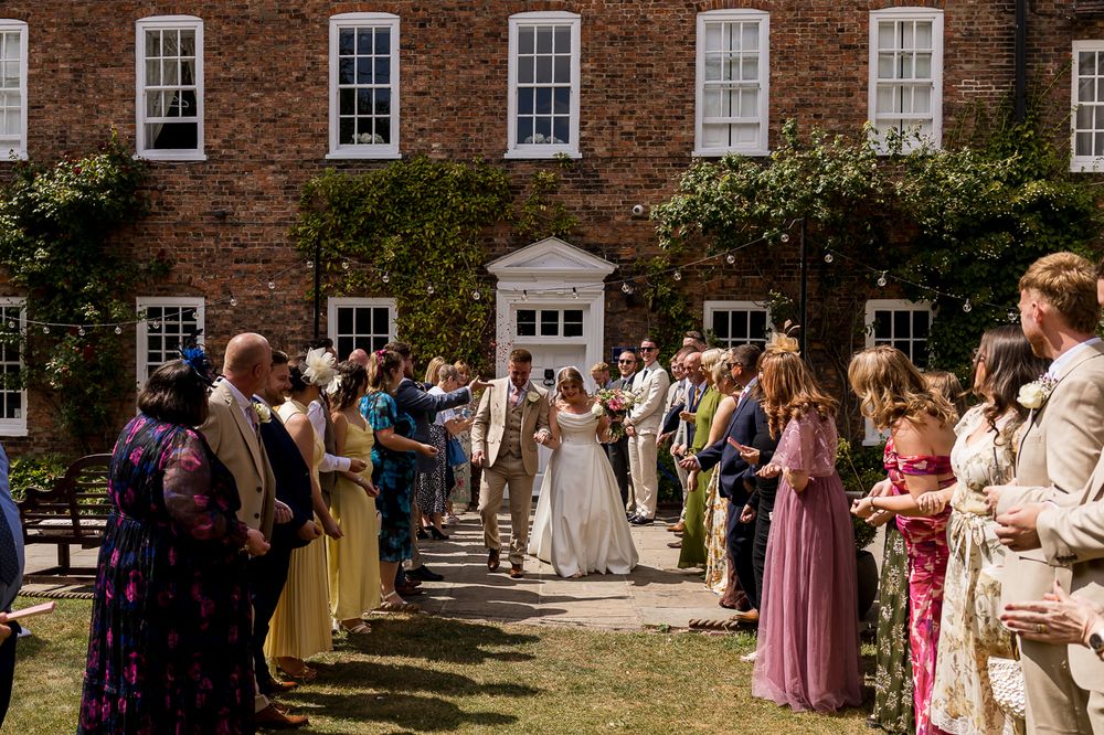 Bride and groom walk through a guard of honor at an outdoor wedding ceremony beside a historic brick manor house.
