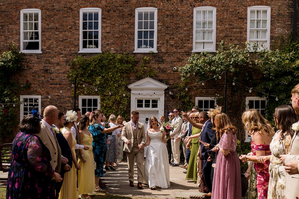 Bride and groom walk through confetti line outside elegant brick wedding venue on sunny day.