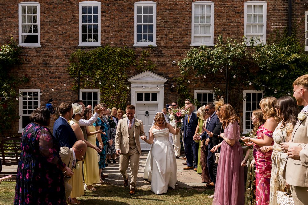 Newlyweds celebrate with guests in confetti tunnel outside a classic Georgian brick manor house.