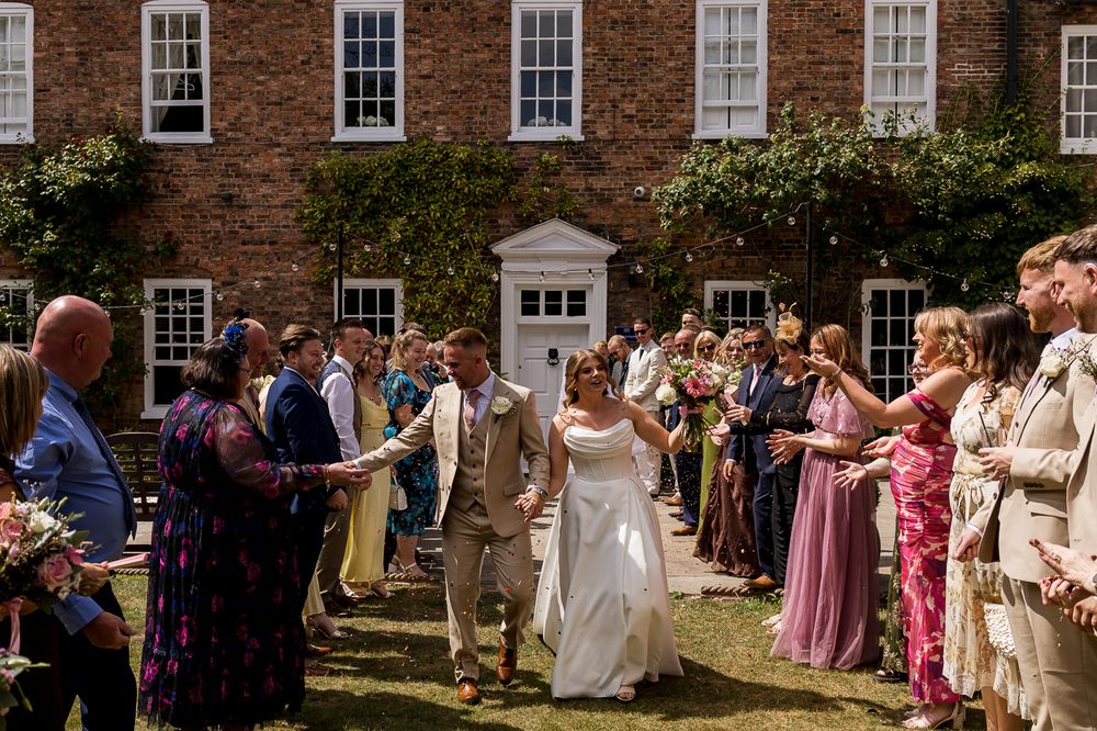 Bride and groom walk through cheering guests outside a brick manor house on a sunny wedding day.