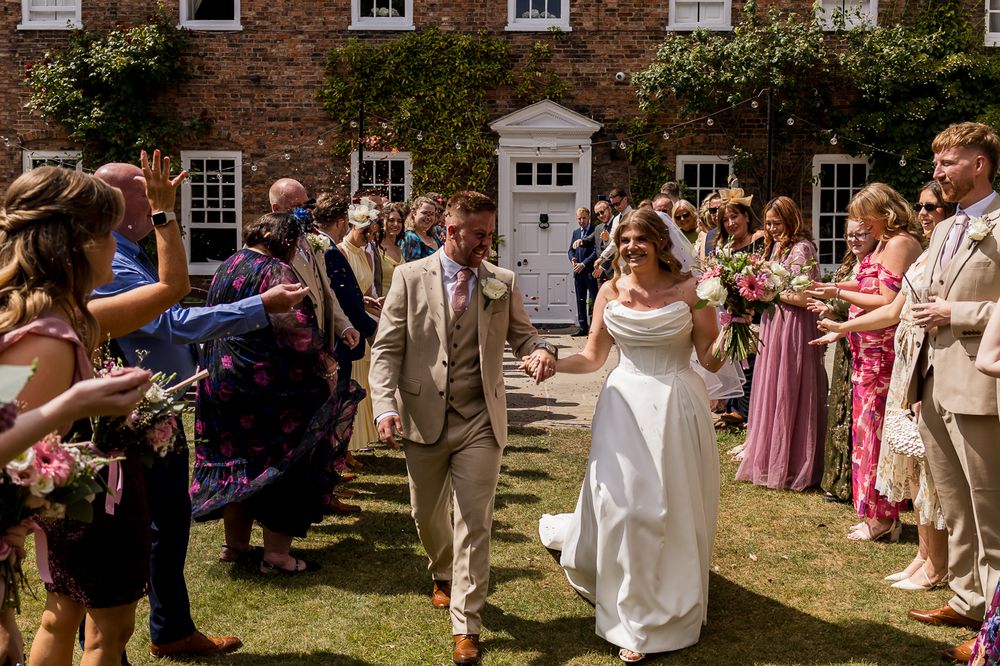 Bride and groom walk through confetti line outside brick manor house on sunny wedding day.