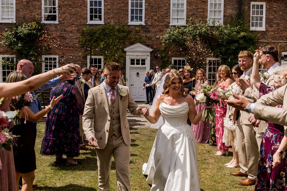 Bride and groom walk through confetti tunnel with smiling wedding guests outside a red brick manor house on a sunny day.