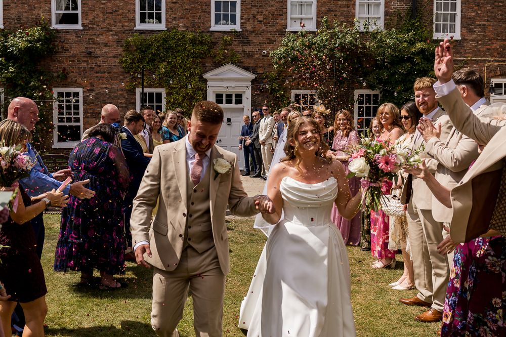 Bride and groom walk through confetti line outside brick manor house on sunny wedding day.