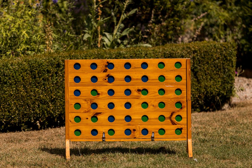 Large wooden Connect Four game board with green and black discs, displayed outdoors on grass with green hedges.