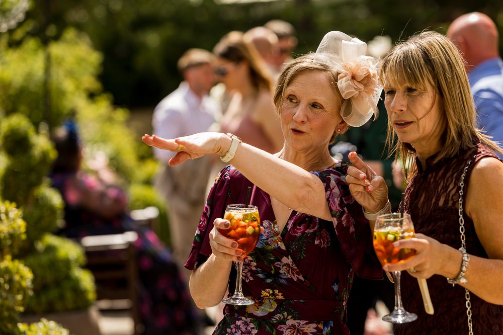 Two elegantly dressed women holding cocktails at an outdoor garden party, one pointing while wearing a fascinator hat.