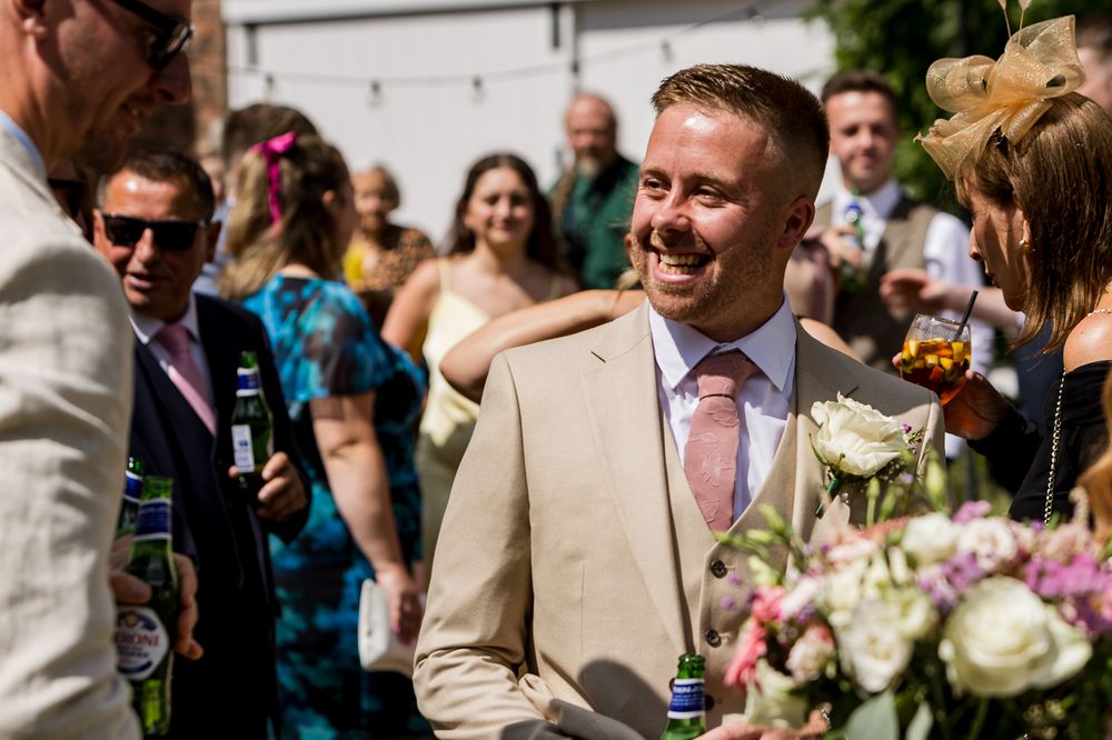Smiling groom in tan suit with pink tie greeting wedding guests outdoors on a sunny day.