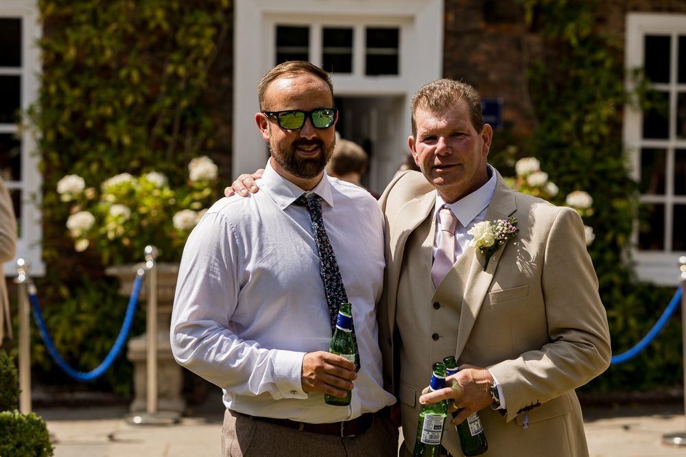 Two men in formal attire smiling outdoors at a wedding, one wearing green sunglasses holding a beer bottle.
