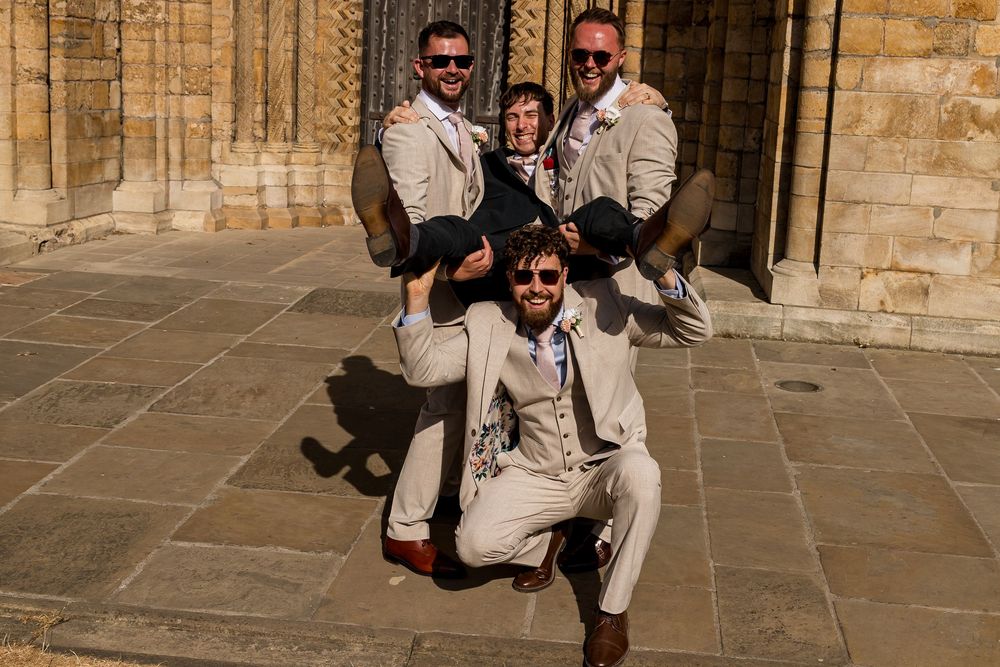 Three groomsmen in tan suits posing playfully outside a stone church on a sunny wedding day.