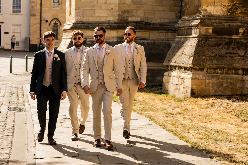 Four groomsmen in suits walking confidently outside a historic stone church on a sunny wedding day.