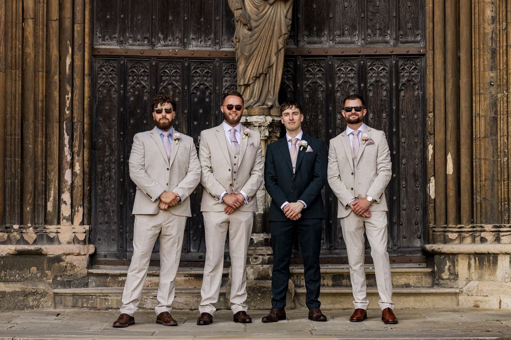 Four groomsmen in suits pose before ornate wooden doors at a wedding venue.