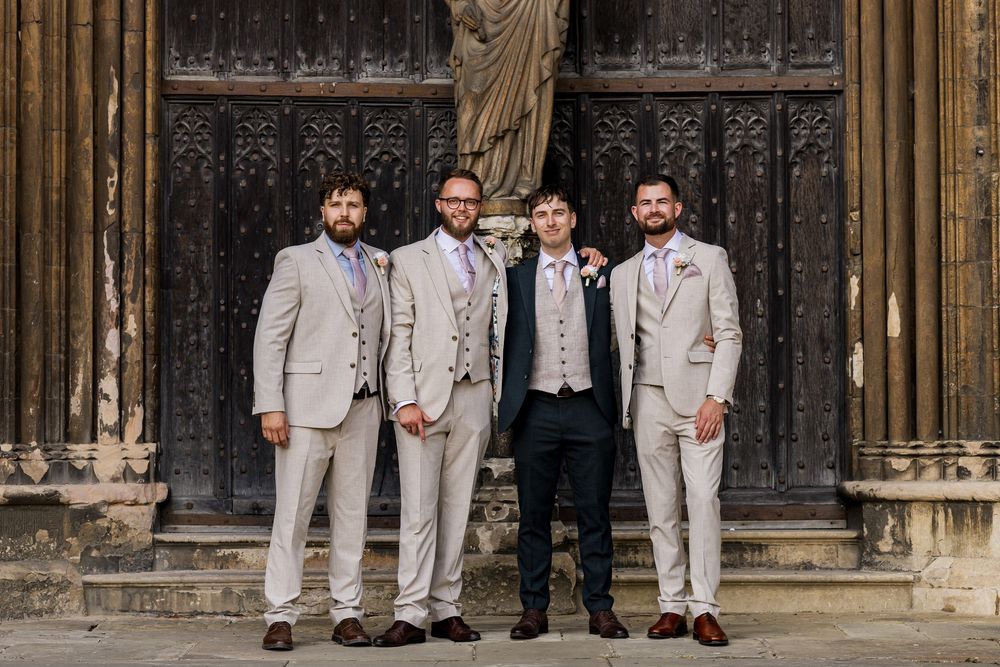 Groom and three groomsmen in beige suits pose before ornate wooden church doors at a wedding.