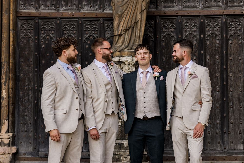 Groom and three groomsmen in matching beige suits pose together in front of ornate wooden doors at a wedding.