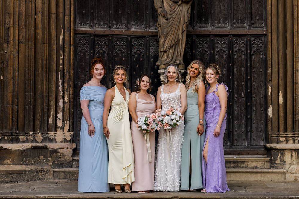 Six bridesmaids in pastel gowns pose with a bride holding a pink bouquet outside a historic stone church.