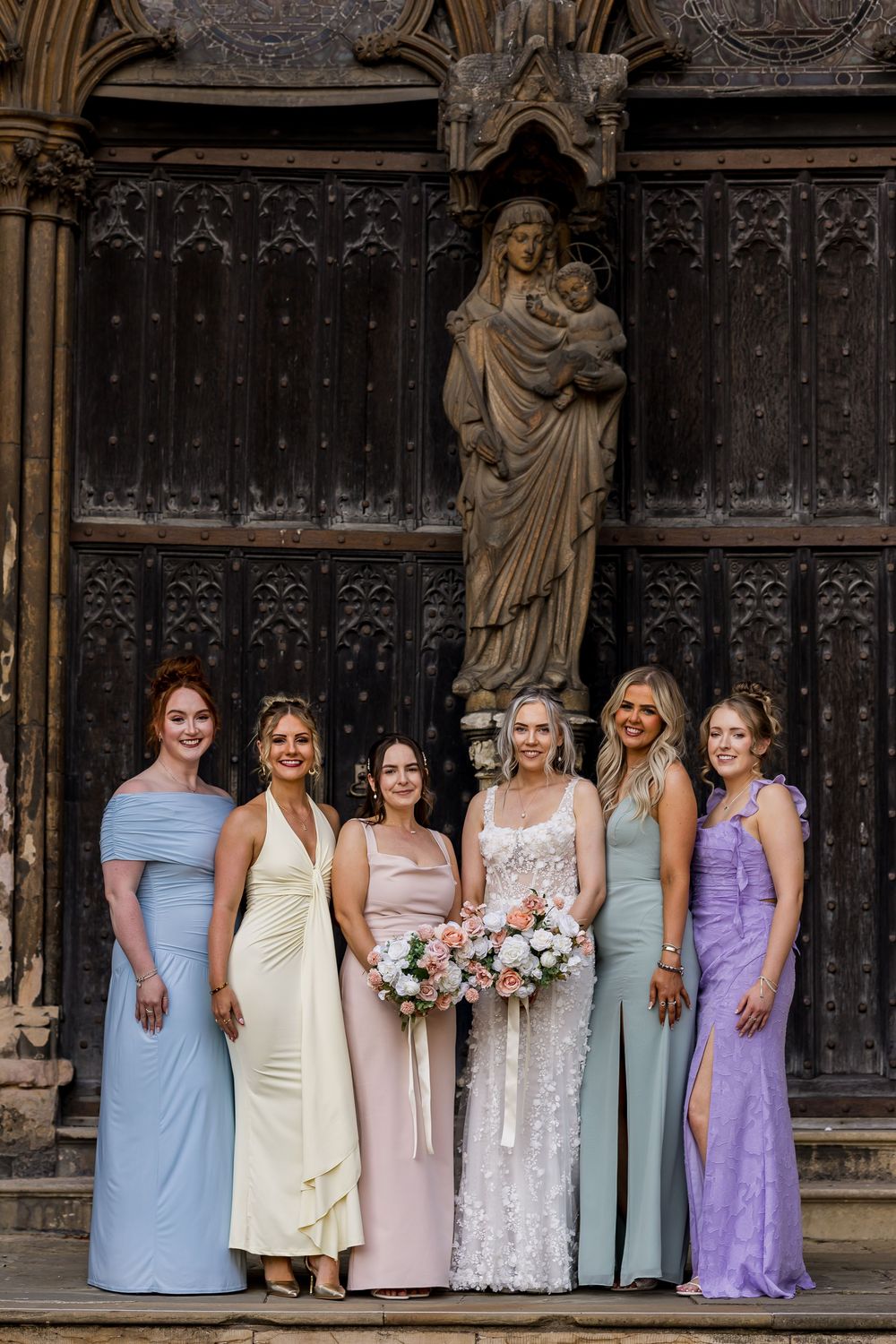 Bride with bridesmaids in colorful gowns posing before ornate wooden doors with stone Madonna statue at wedding.