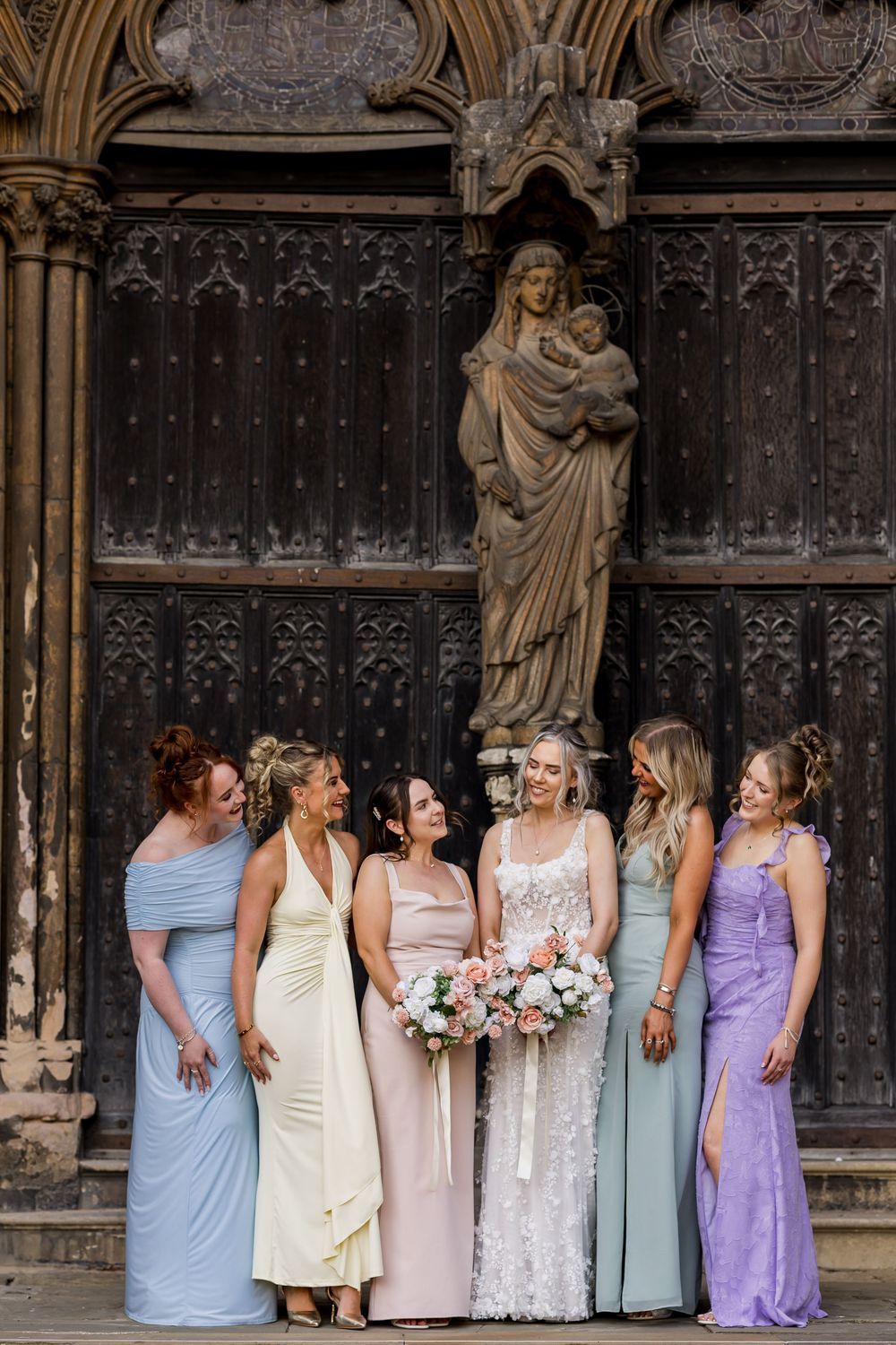 Bride with bridesmaids in pastel gowns holding bouquet before ornate gothic church doors with stone Madonna statue.