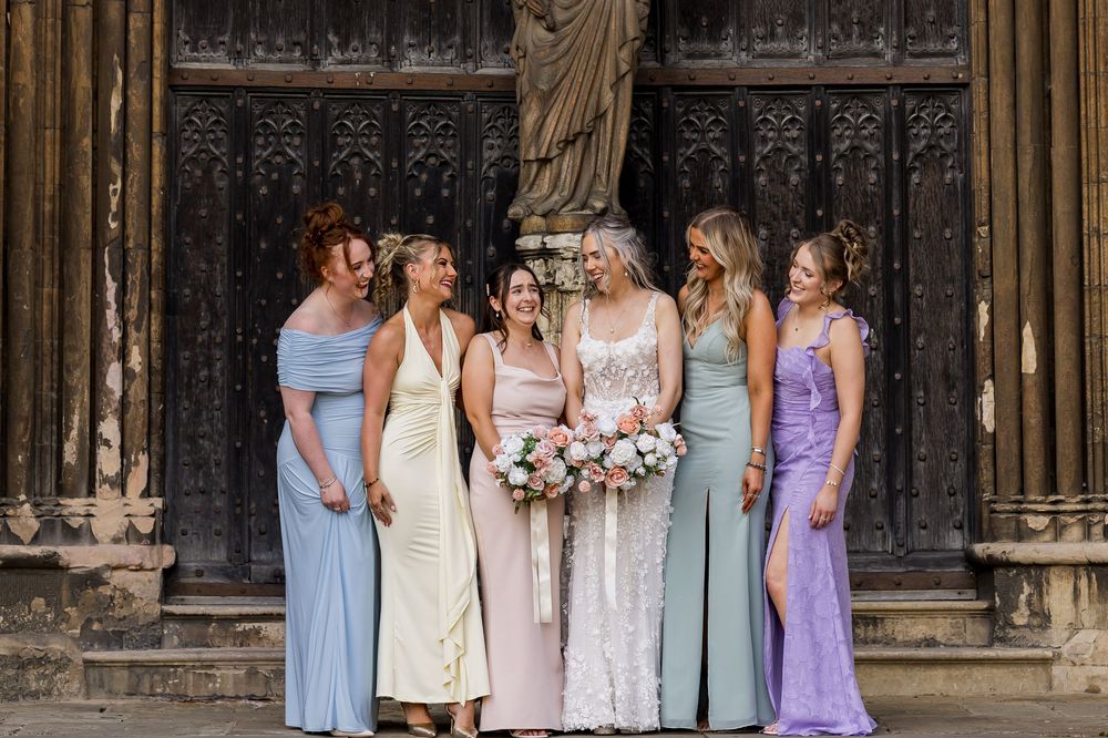 Bride in lace gown holds bouquet surrounded by bridesmaids in pastel dresses outside ornate wooden church doors.