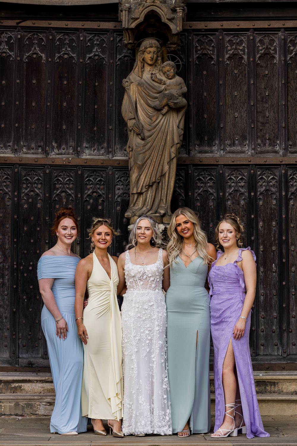 Bride in white floral gown poses with four bridesmaids in pastel dresses before ornate wooden church doors.