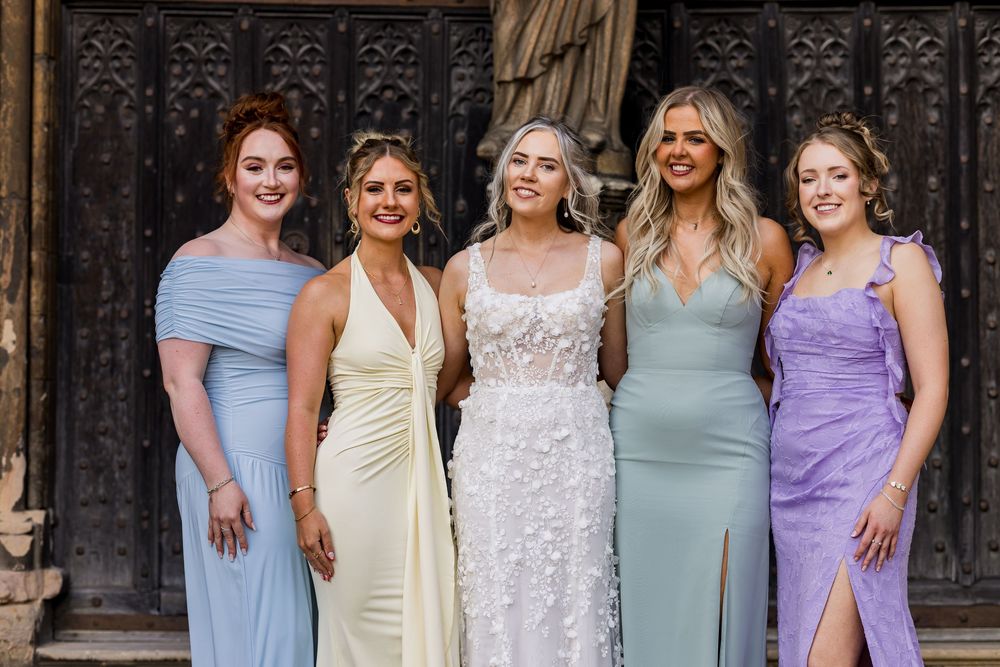 Bride in floral gown poses with four bridesmaids in pastel dresses outside ornate wooden doors.