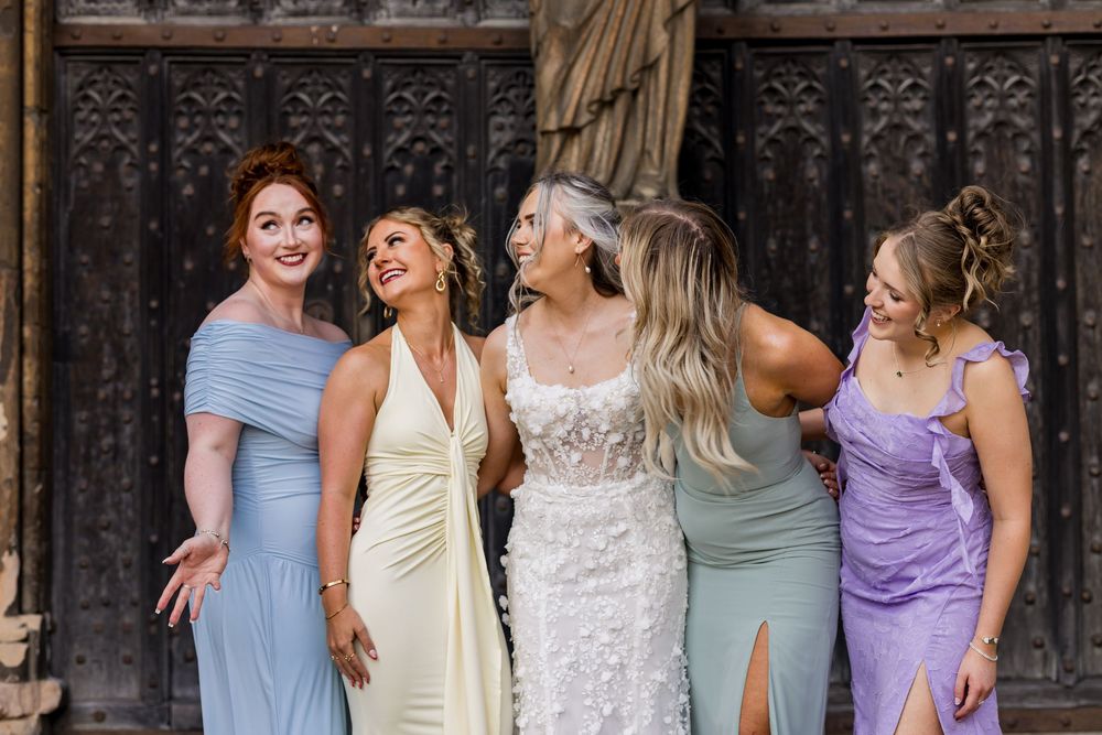 Bride in white floral gown laughing with four bridesmaids in pastel dresses outside ornate wooden doors.