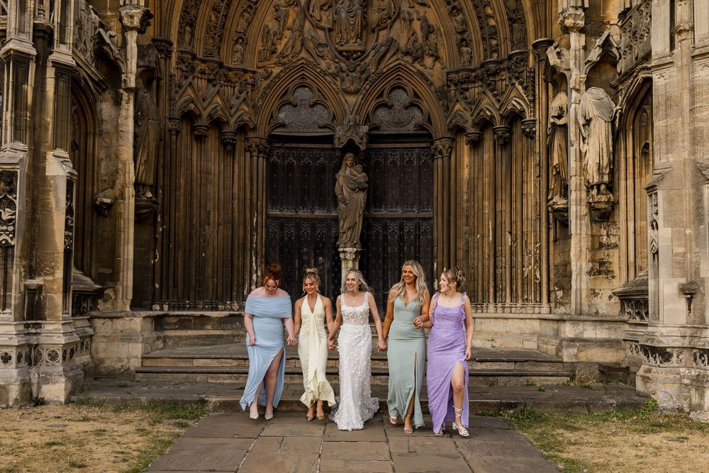Bride and four bridesmaids in colorful dresses pose before ornate Gothic cathedral doors on a sunny wedding day.