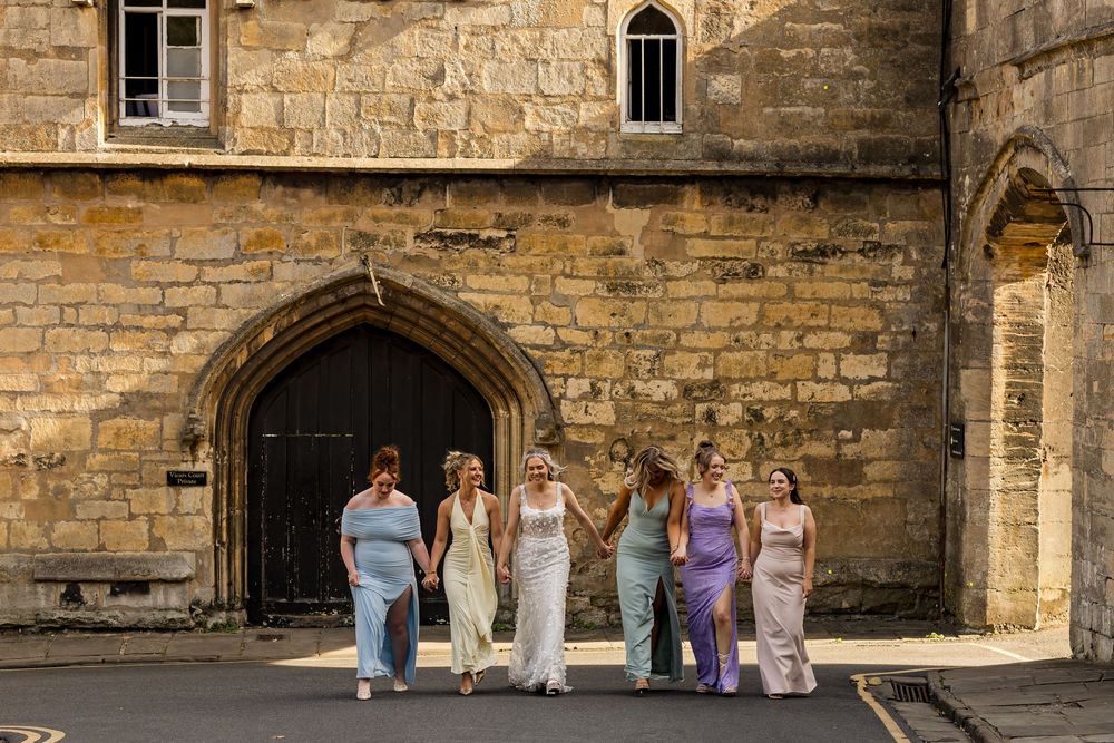 Bride and bridesmaids in pastel gowns walking together outside a historic stone building on a sunny day.