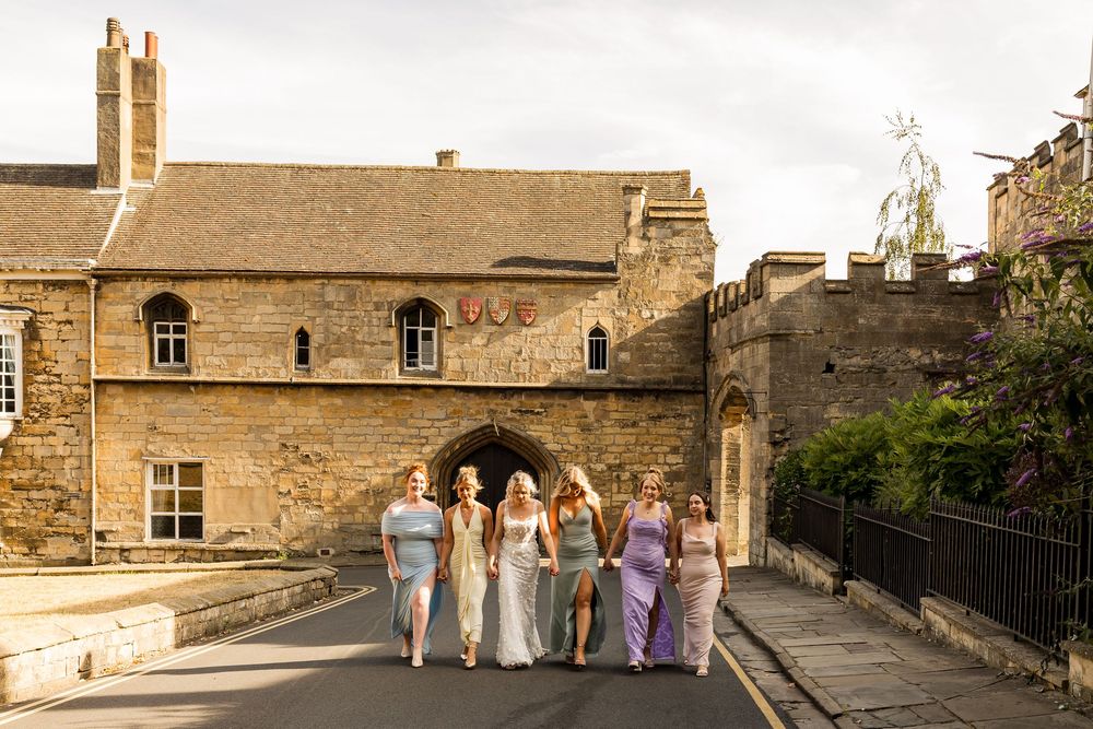 Six bridesmaids in pastel dresses walk together outside a historic stone building on a sunny day.