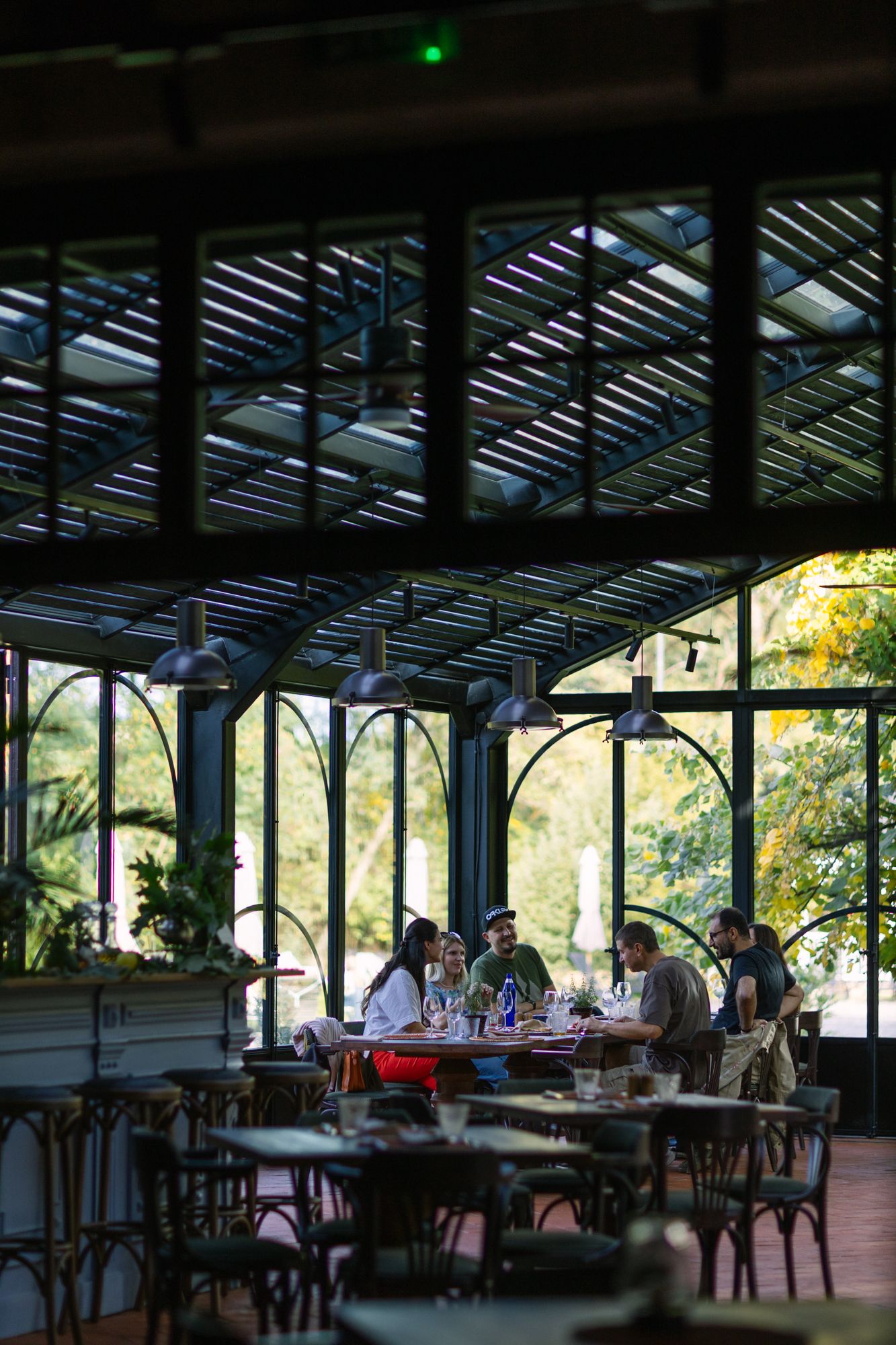 Group of people dining in an elegant glass greenhouse restaurant surrounded by lush greenery.
