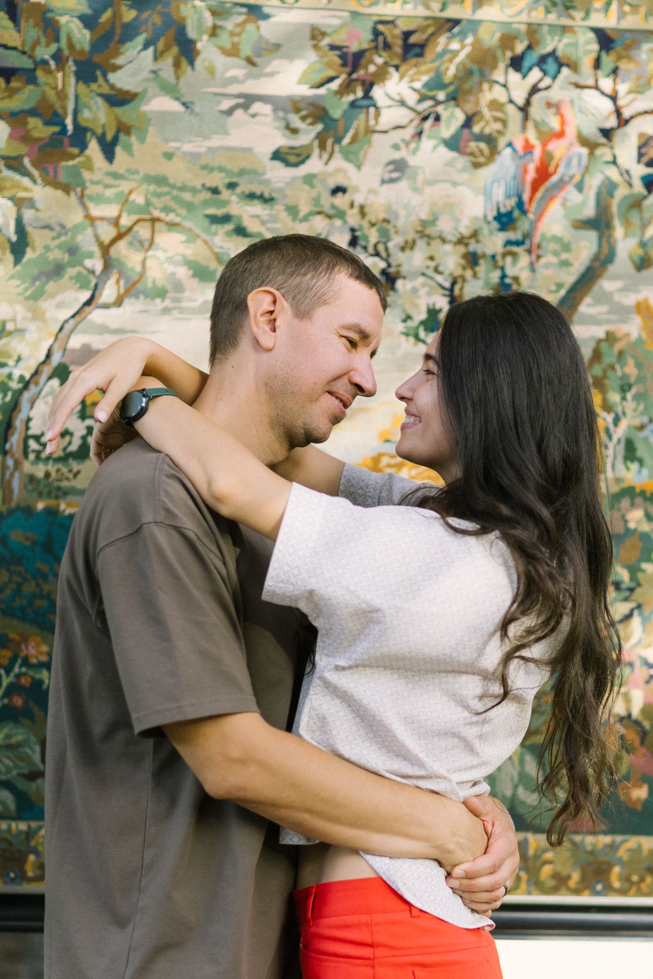 A couple embraces and smiles at each other in front of a colorful floral tapestry wall hanging.