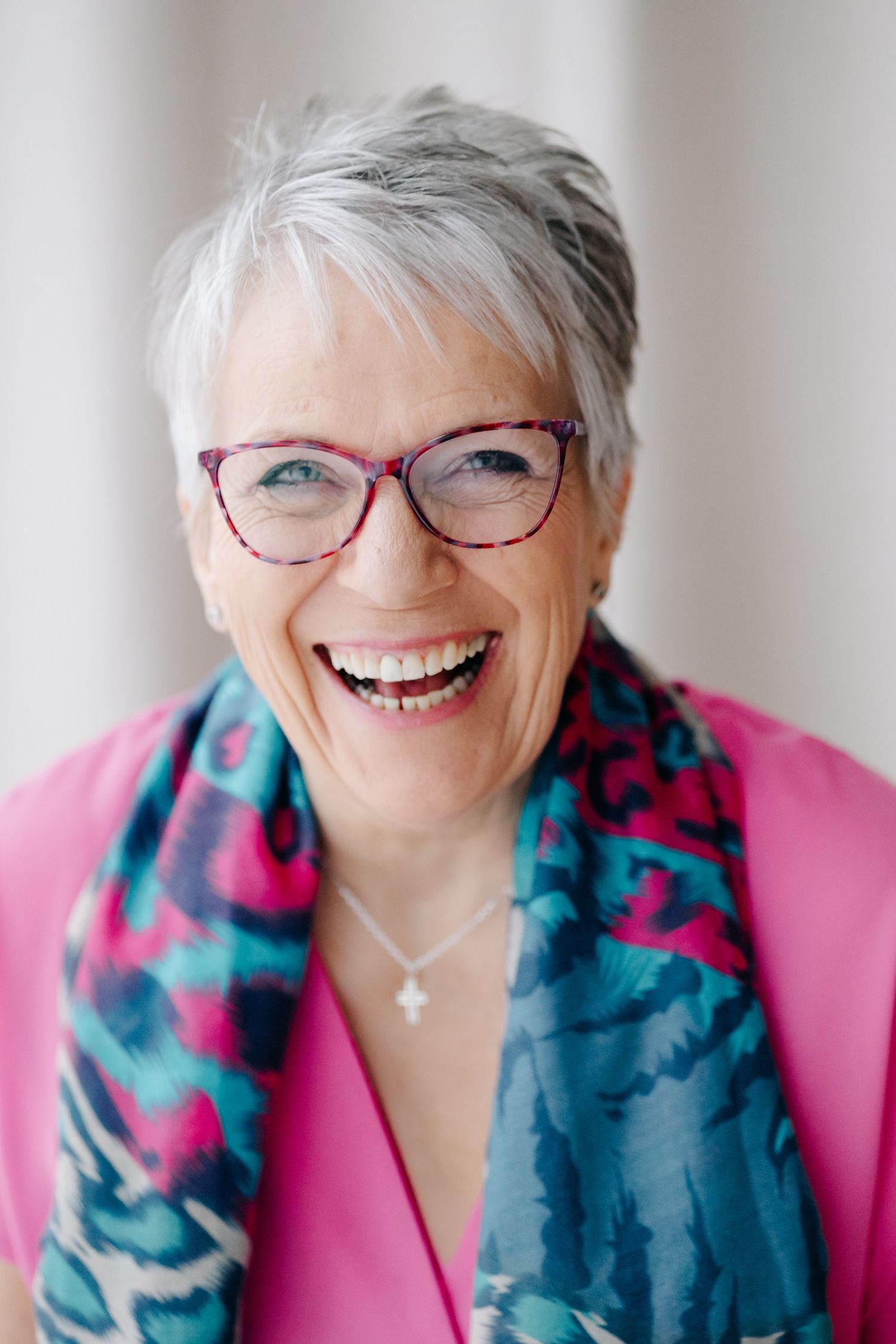 Smiling older woman with short gray hair, pink top, colorful scarf, and purple glasses laughing joyfully.