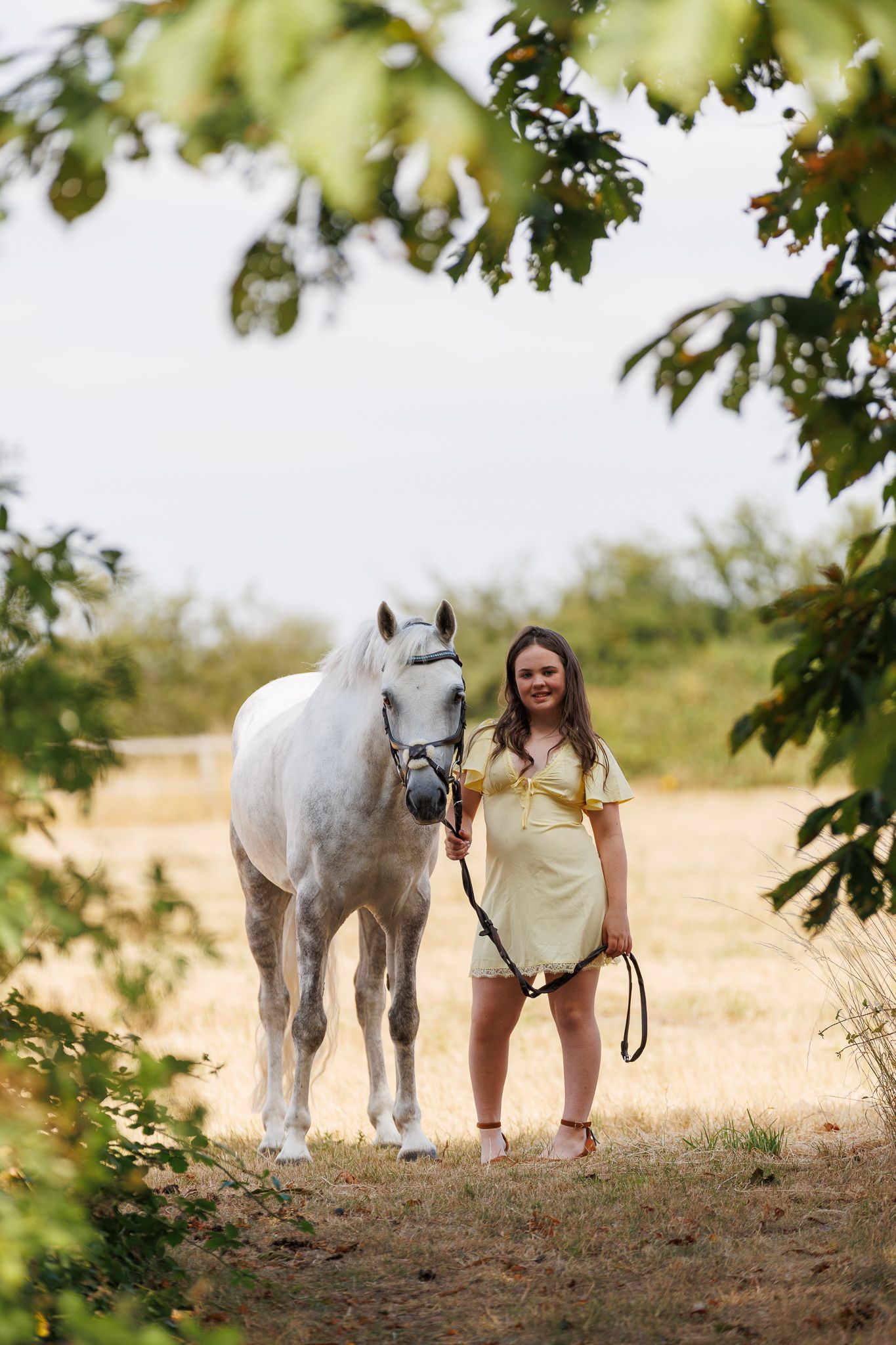 Young woman smiling between two bay horses with white markings, holding reins near a brick stable.