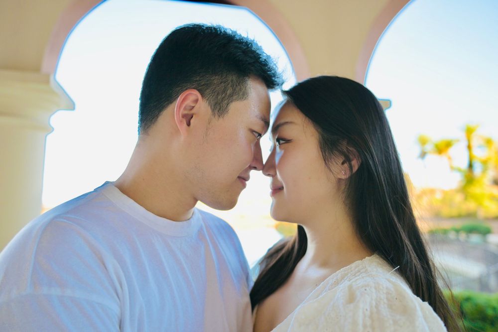 Romantic couple touching noses in a tender moment beneath a sunlit archway, with soft golden light and greenery in the background.