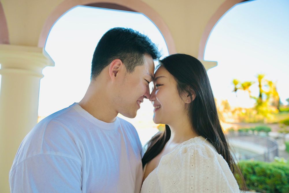 Romantic couple touching foreheads and smiling softly under a sunlit stone archway with a blurred garden in the background.