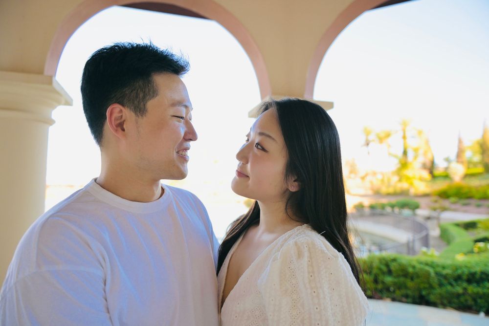 A smiling couple gazes at each other under a stone archway with a sunlit garden and soft golden light in the background.