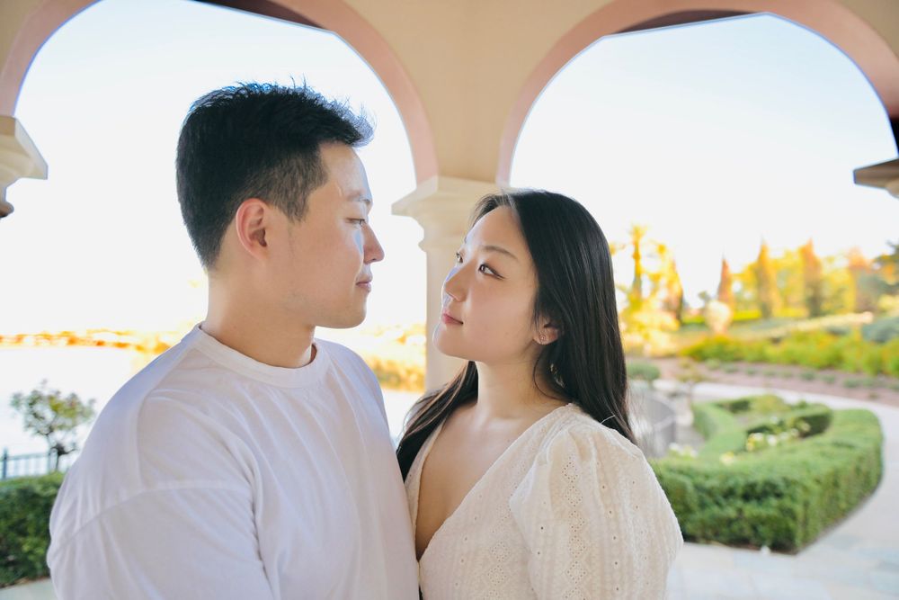 Couple in white outfits stand face to face under a sunlit stone archway with a scenic garden and water view behind them.