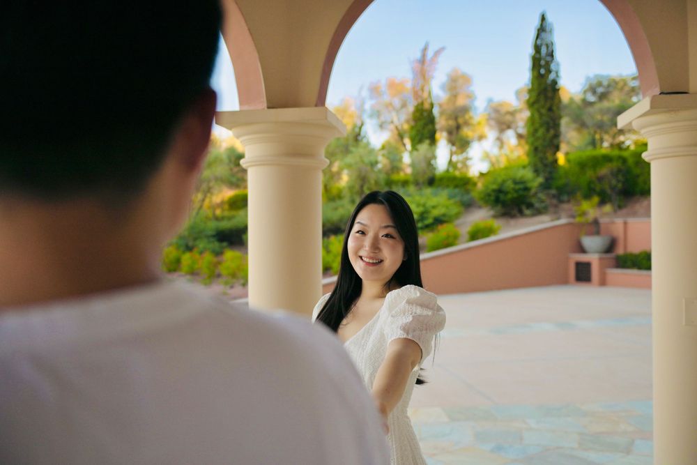 Smiling woman in a white dress stands under a sunlit stone archway, framed by a blurred figure in the foreground and lush garden greenery.