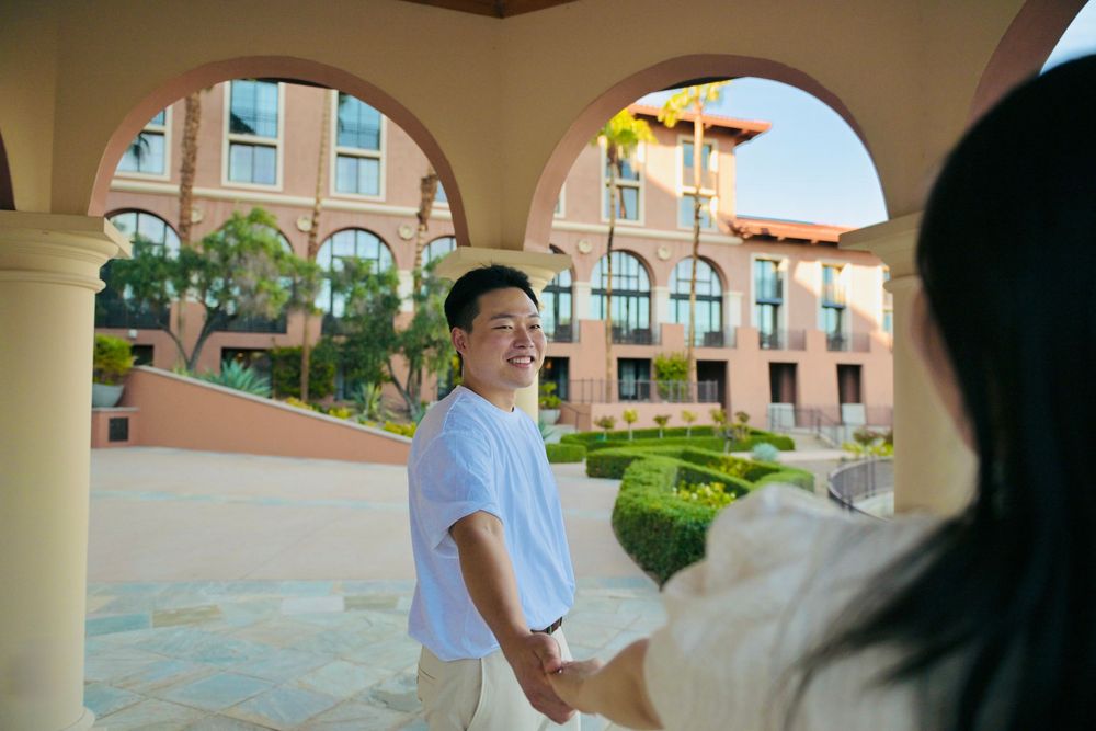 Smiling man in a light blue shirt holds hands with a woman under a sunlit stone archway at a resort with terracotta buildings.