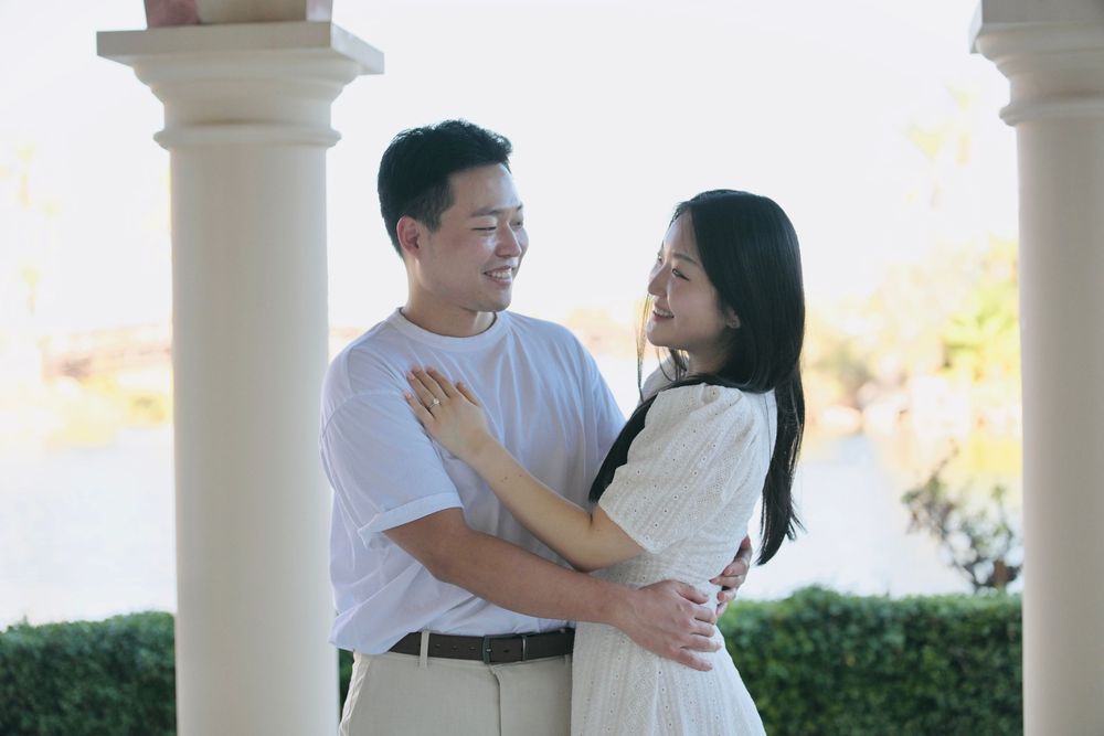 A smiling couple embraces between white columns on a covered terrace, with a softly blurred waterfront and green hedge behind them.