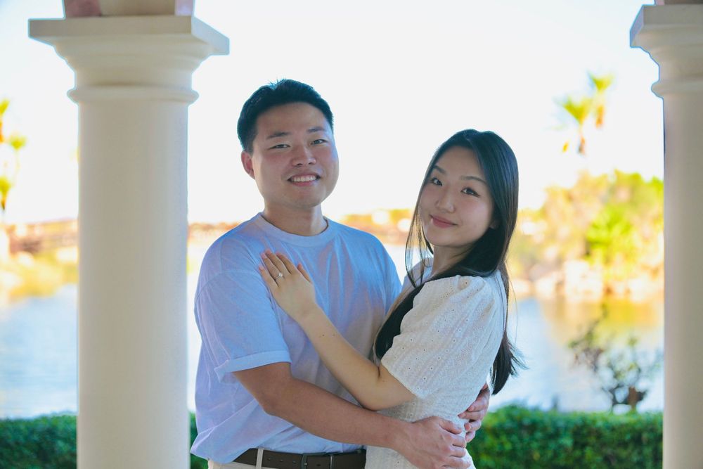 Smiling couple embracing between white columns with a scenic lake and palm trees in the background.