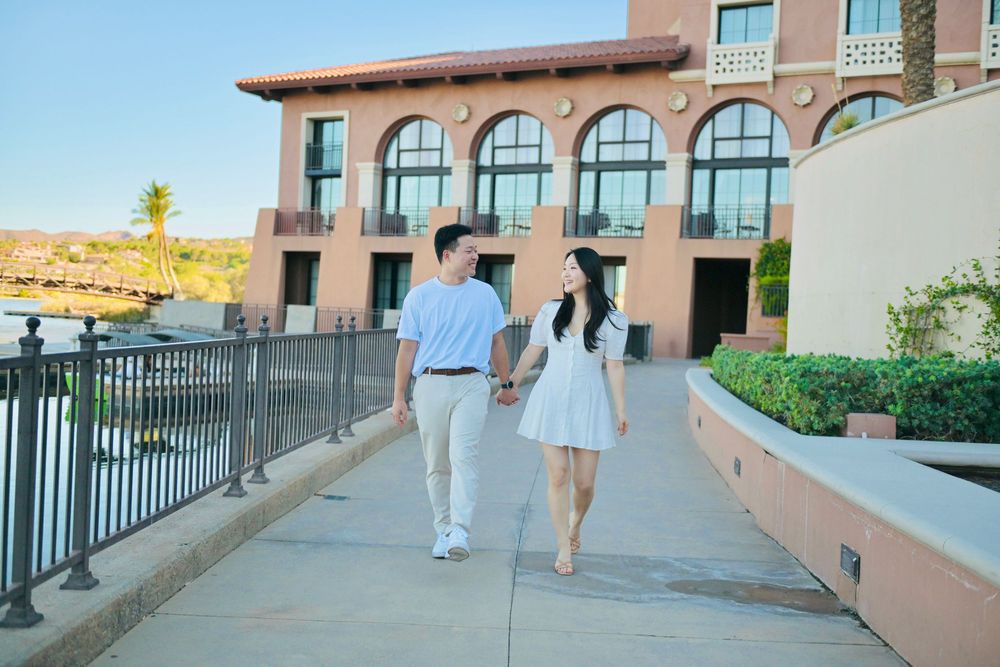 Couple holding hands walks along a paved resort path past a terracotta hotel, palm trees and manicured hedges under a clear blue sky.