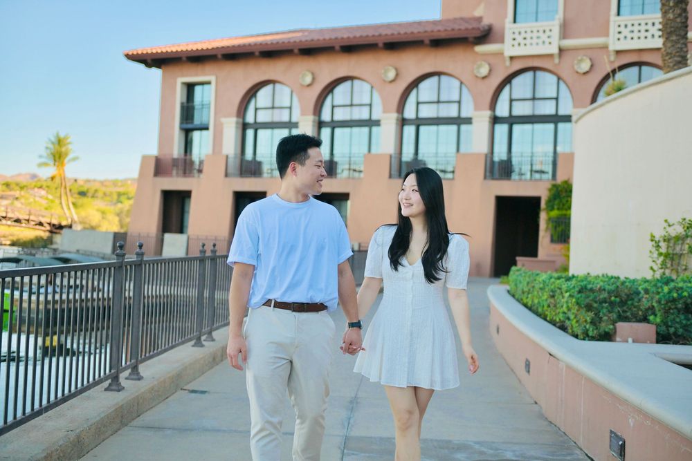 Couple in matching light outfits holding hands while walking along a sunny path near a terracotta-colored arched building.