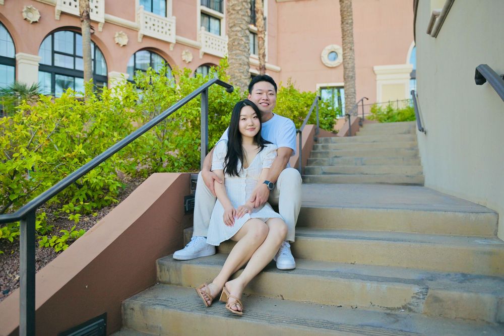 Smiling couple in white outfits sitting close together on outdoor stone steps beside a green hedge and warm-toned building.
