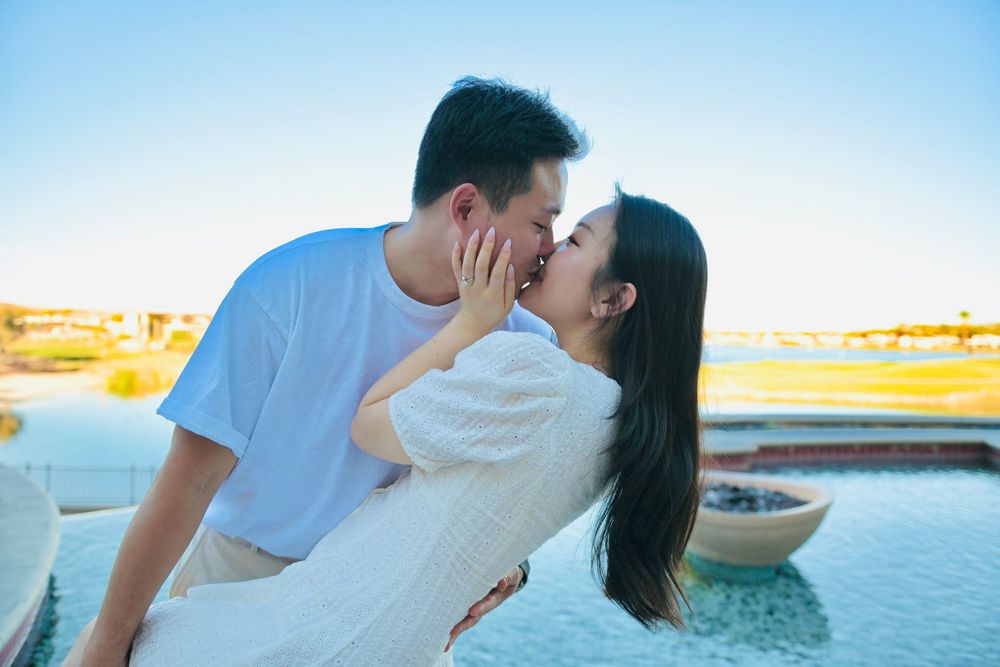 Romantic couple kissing beside a resort infinity pool at sunset, the woman in a white dress and the man in a light blue t-shirt.