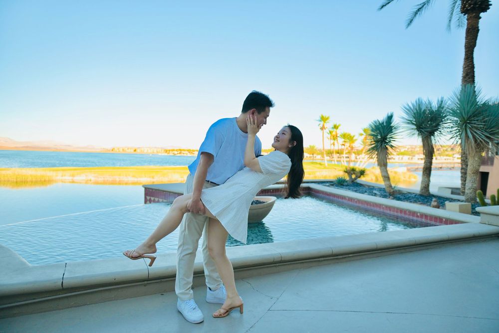 Couple in a romantic dip pose beside a resort pool with palm trees and a scenic lake view under a clear blue sky.