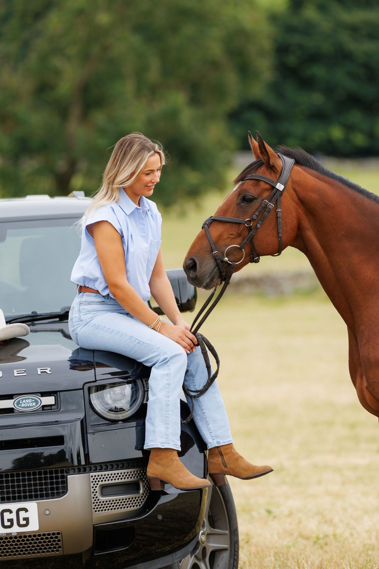 Blonde woman in a blue shirt and jeans sits on the front of a Land Rover Defender, smiling at a brown bridled horse in a sunny field.