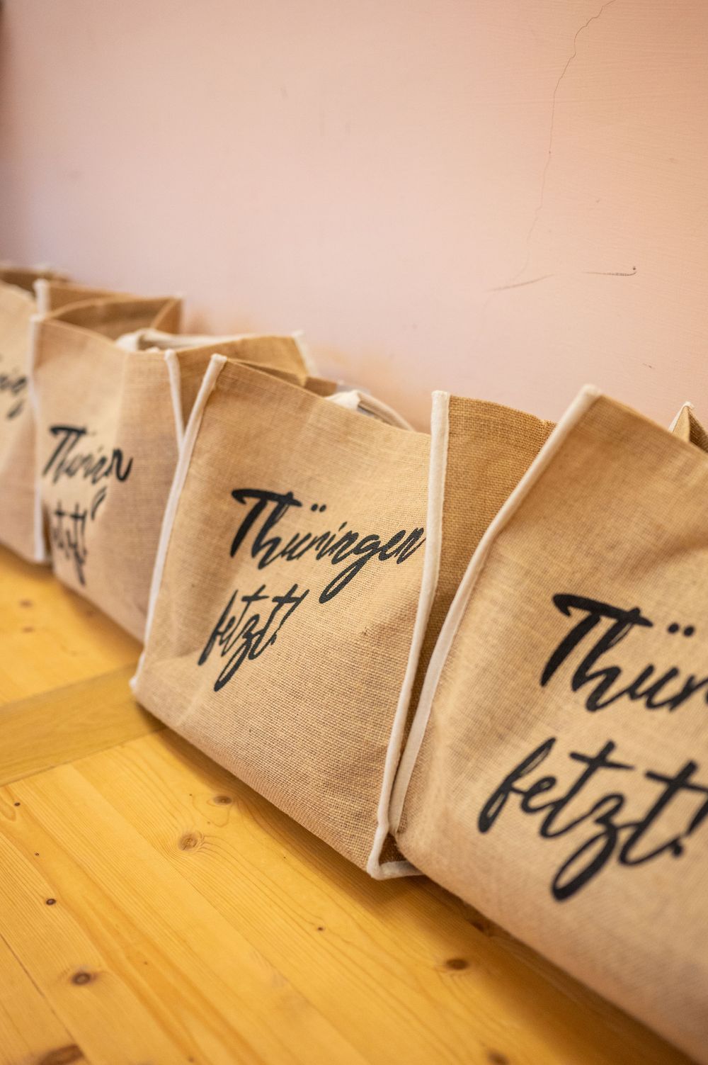 Row of natural jute tote bags printed with the handwritten slogan Thüringen fetzt lined up on a light wooden floor against a pink wall.