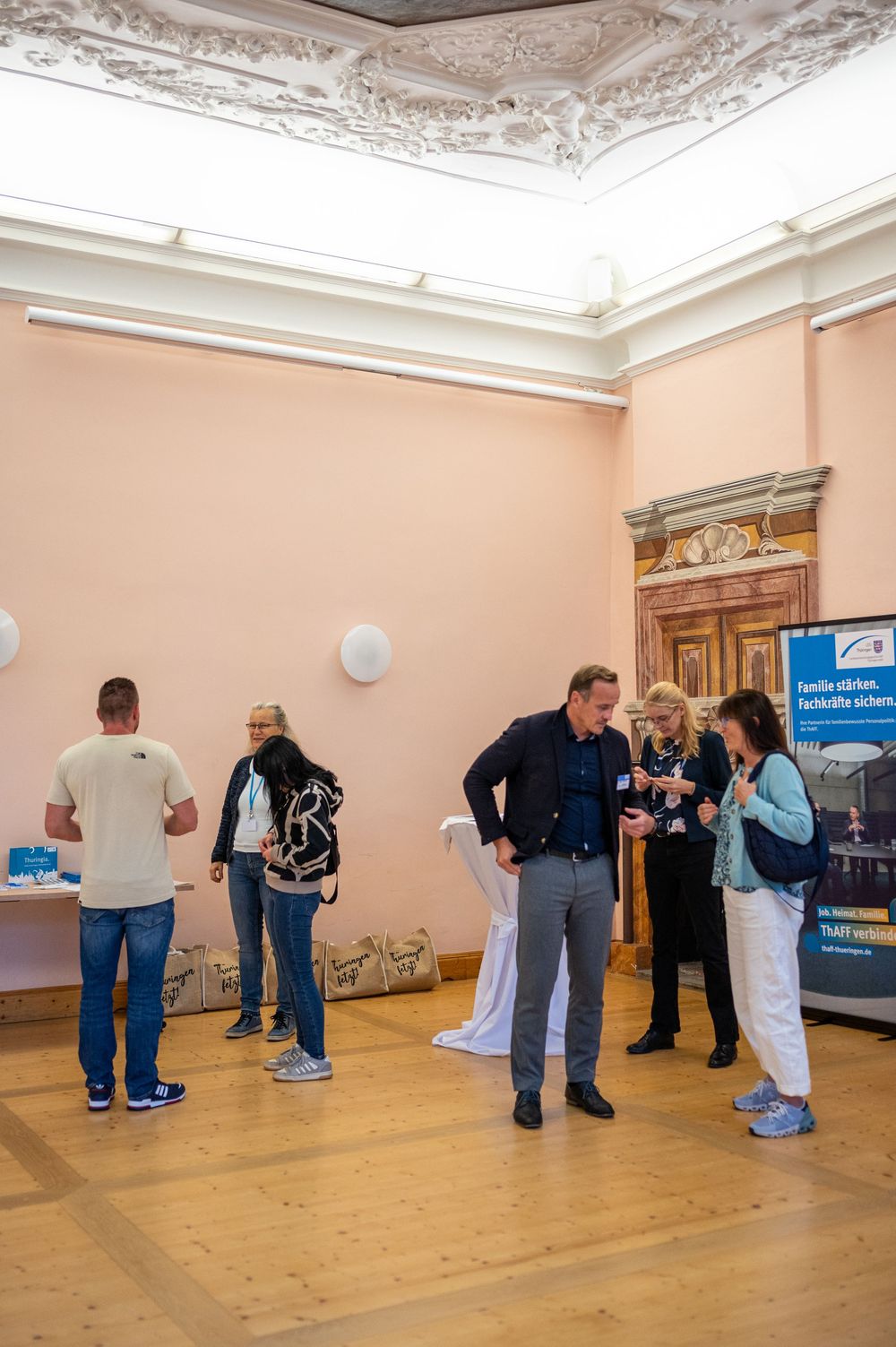 Guests mingle and chat in an ornate event hall with stucco ceiling, pink walls and parquet floor beside a banner and information table.