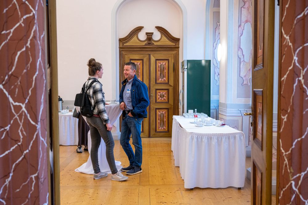 Two people stand talking in an elegant hall with ornate wooden doors, parquet flooring, and a white-clothed table nearby.