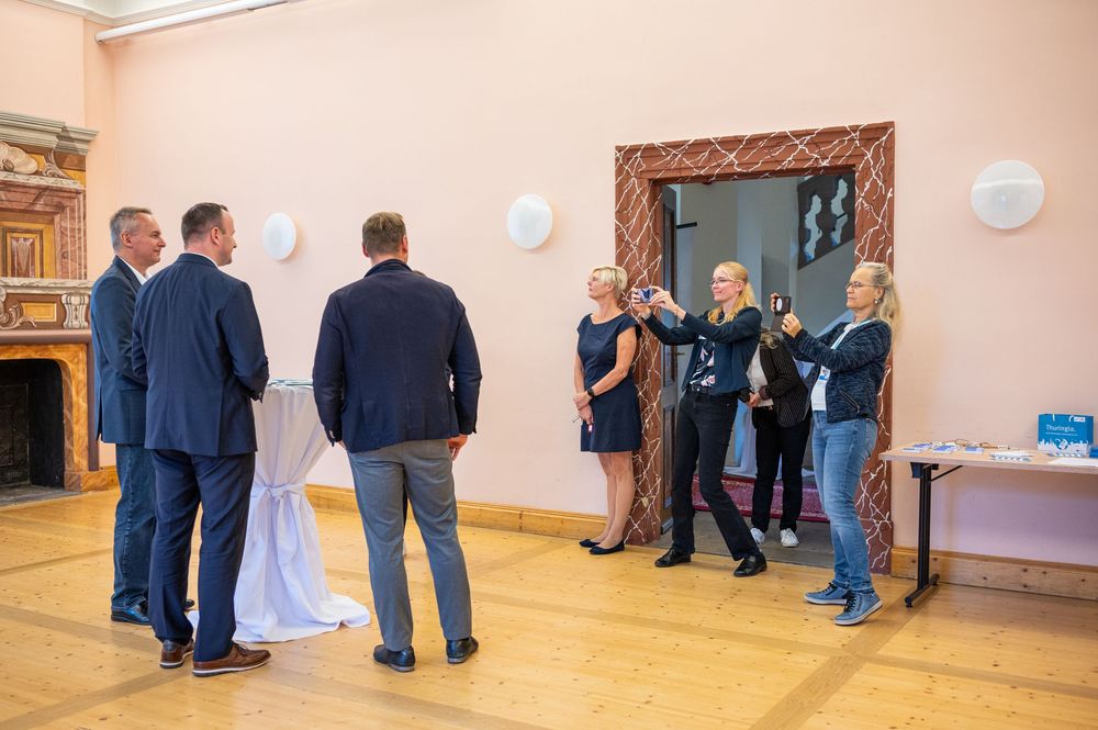 Three men in dark blazers stand beside a draped cocktail table while three women photograph the scene in a pink hall with wood flooring.