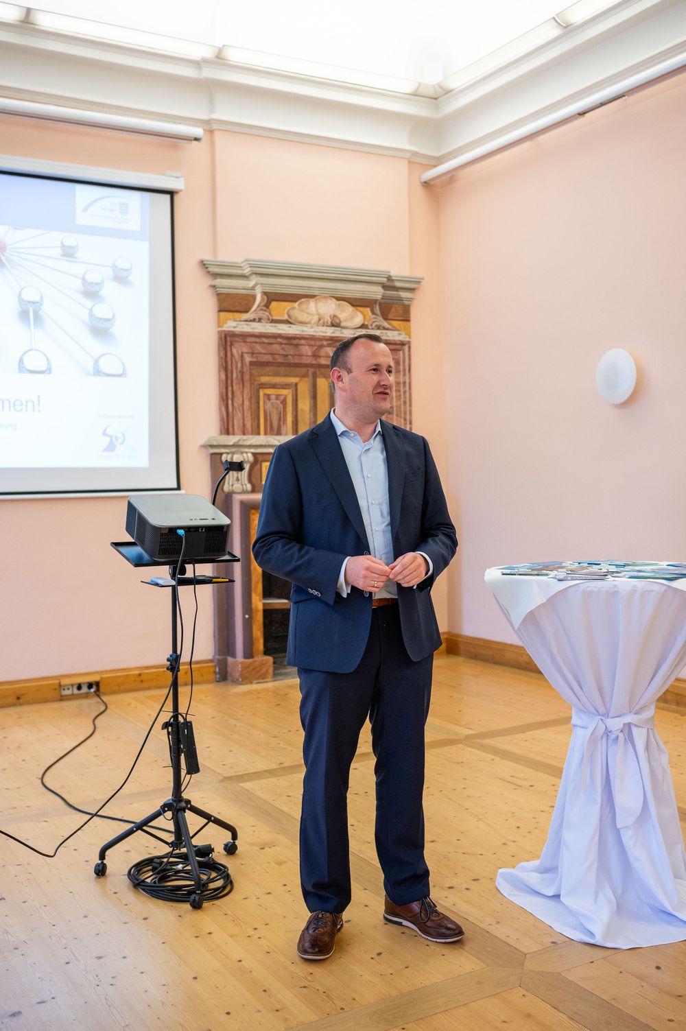 Man in a navy blazer presents in an ornate room with pink walls, a projector and screen beside him, and a draped cocktail table.