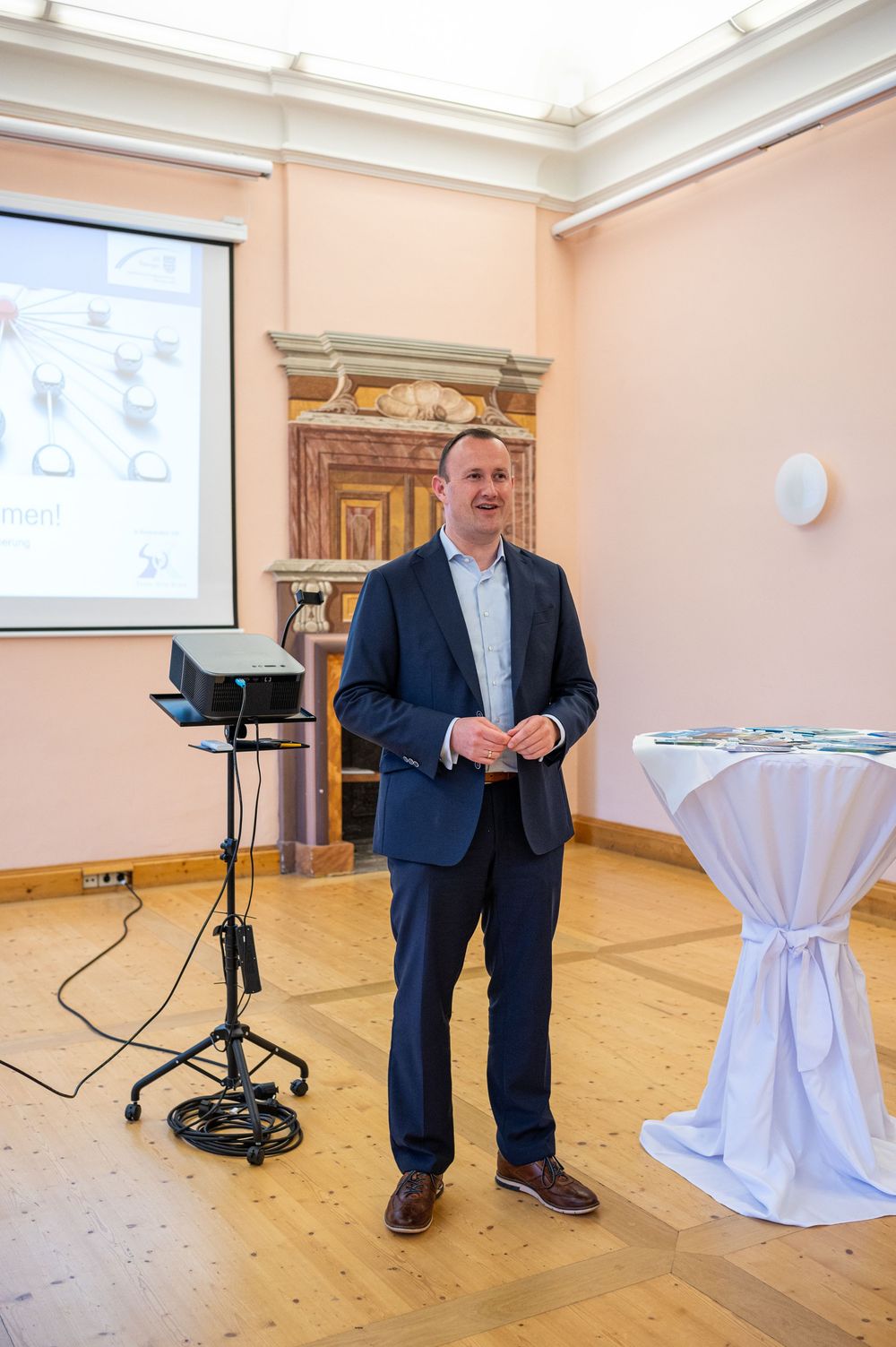 A man in a navy blazer and blue shirt speaks during a presentation in an elegant hall with a projector, screen and draped standing table.